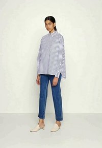 Prevelika bijela košulja s mornarskim verticalnim prugama, uparena s plavim ravnim trapericama i bež slip-on cipelama. Jednostavan dizajn, ležeran kroj.