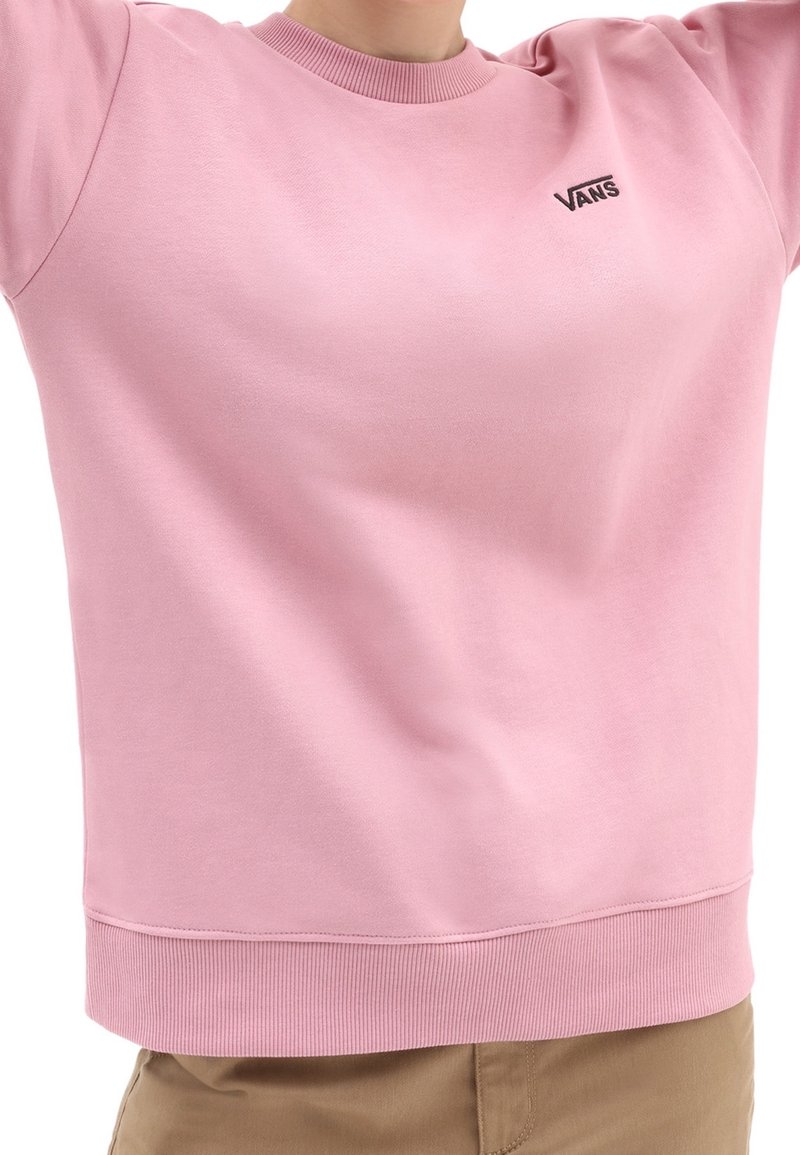 Personne portant un sweat-shirt Vans rose clair avec col et ourlet côtelés, associé à un pantalon beige, les bras levés hors du cadre.