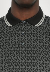 Polo shirt noir avec un motif de logo gris répétitif, un col noir avec des rayures grises, et une patte de boutonnage à trois boutons avec des boutons brillants.