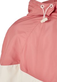 Lekka, wodoodporna kurtka z przeważającą różową górną częścią i białą dolną panelem, z regulowanym kapturem na sznurku i gładką teksturą.