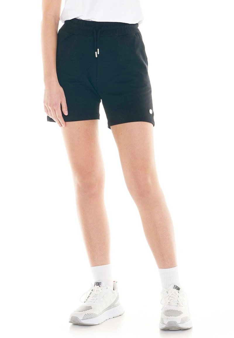 Un mannequin porte un short de sport noir à cordon, un haut blanc, des chaussettes blanches et des chaussures de course blanches sur un fond blanc uni.