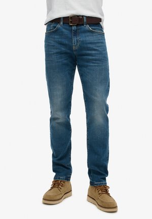 Blue denim jeans with a relaxed fit, faded details, five-pocket design, and a brown belt. Tan suede shoes complete the look.