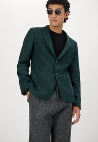 Mørkegrønn ullblandingsblazer med en knapp lukking, notched lapels, to frontlommer og en skreddersydd passform. Båret over en svart topp.