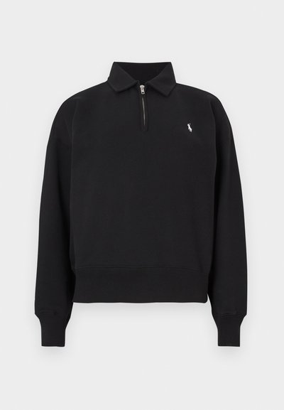 Svart genser med halv glidelås, krage, lange ermer og ribbestrikkede mansjetter. Har en liten hvit logo på venstre bryst.