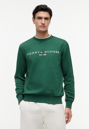 Άνδρας με πράσινο φούτερ Tommy Hilfiger με το λογότυπο και άσπρο παντελόνι, σε απλό φόντο.