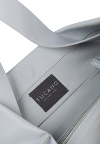 Interno di una borsa tote in pelle grigia chiara con etichetta in tessuto scuro recante la scritta "Tucano Milano 1985", tasca con zip e cuciture lisce e a contrasto.