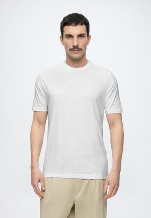 Homme aux cheveux foncés et moustache, portant un t-shirt blanc BOSS et un pantalon beige, debout devant un fond clair uni.