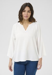 Vit långärmad blus med V-ringning, en strukturerad yta och avslappnad passform. Kanske ihop med blå denimjeans, vilket ger en bekväm stil.