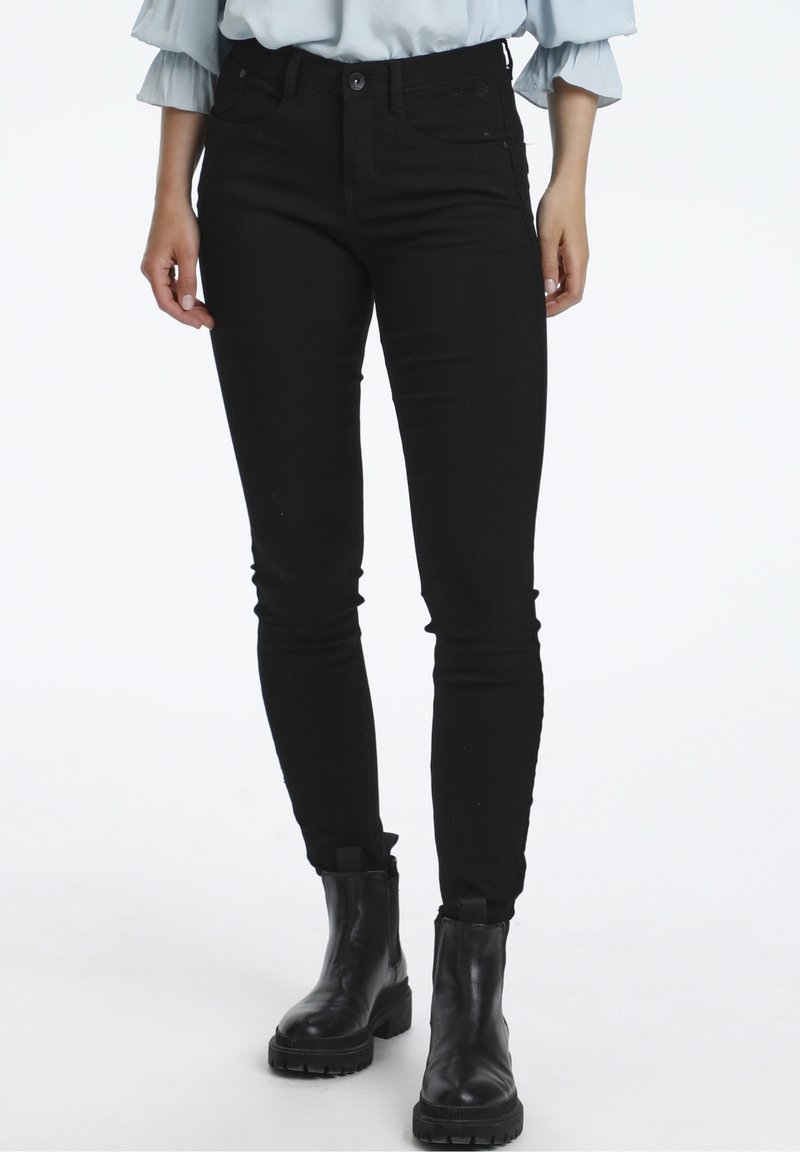 Femme portant un pantalon noir ajusté et des bottines noires épaisses, se tenant contre un fond blanc.