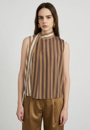Frau trägt eine ärmellose, braun-beige vertikal gestreifte Bluse mit Schaldetail und passenden braunen Satin-Hosen, stehende vor einem einfarbigen Hintergrund.