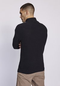 Suéter negro de punto acanalado con un cuello alto, que presenta un acabado texturizado y un dobladillo curvado, combinado con pantalones beige claros.