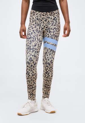 Osoba nosiaca béžovo-čierne legíny s dvomi svetlomodrými pásikmi na stehne a bielu športovú obuv.