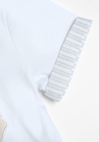 Camisa de manga corta azul claro con un puño a rayas pálido y una superficie texturizada. Presenta un gráfico detallado en tonos neutros en el frente.