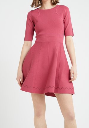 Mujer con un vestido rosa ajustado con mangas hasta el codo y una falda acampanada con detalle de dobladillo en zigzag.