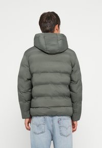 Πληρωμένη ελαιόχρωμη ζακέτα με κουκούλα, με ψηφιακή υφή και τετράγωνη σιλουέτα, φορεμένη πάνω από ανοιχτόχρωμα μπλε τζιν.