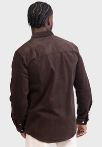 Brązowa koszula z sztruksu z kołnierzykiem, długimi rękawami i zapięciem na guziki przy mankietach. Posiada gładką fakturę i zaokrąglony dół z tyłu.