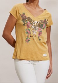 Una camiseta amarilla con cuello en V y estampado de cráneo de toro decorado con flores, combinada con pantalones blancos, vista de los hombros a las caderas.