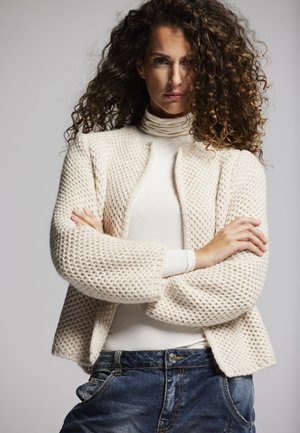 Strickcreme-Cardigan mit strukturiertem Muster, offener Front und weiten Ärmeln, kombiniert mit einem weißen Rollkragenpullover und blauen Jeans.