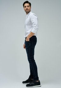 Camisa blanca de botones con mangas enrolladas, jeans ajustados azul oscuro y zapatillas deportivas negras con suelas blancas. Fondo neutro.