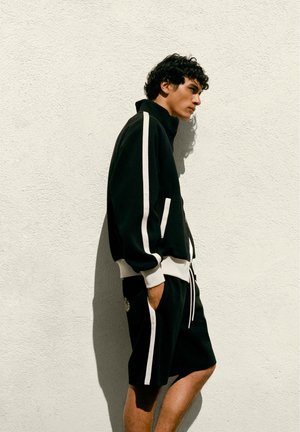 Fiatal férfi fekete sportdzsekiben és fehér csíkos rövidnadrágban, oldalra fordulva egy texturált fehér fal előtt áll, kezei a zsebében.