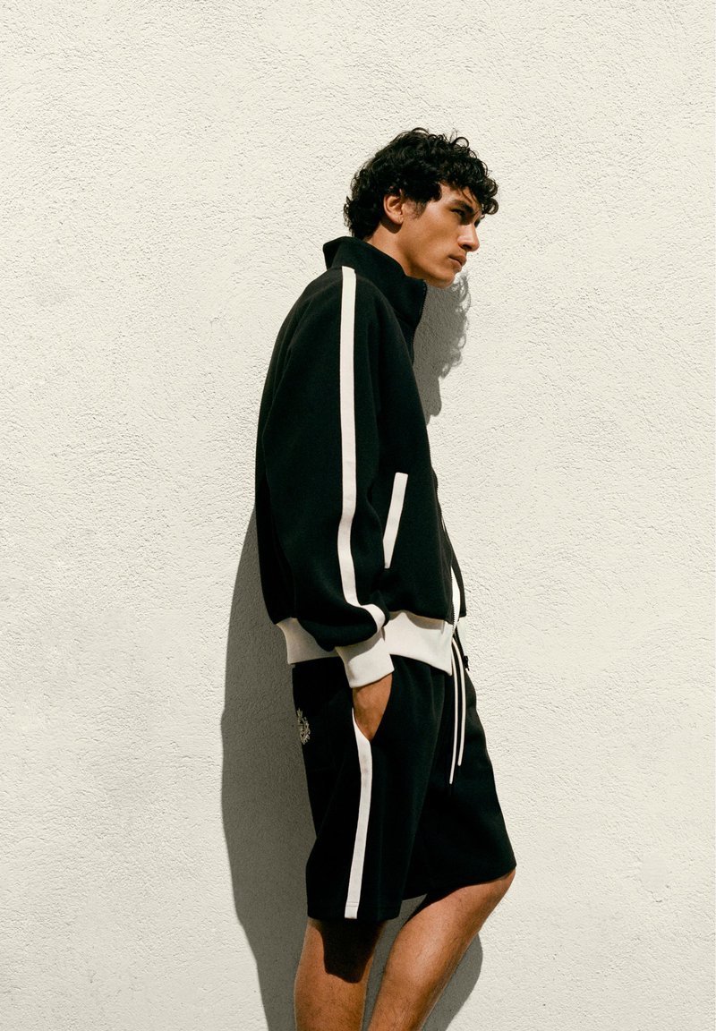 Jeune homme en veste de sport noire et short à rayures blanches, debout de profil contre un mur blanc texturé, les mains dans les poches.