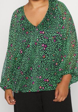 Persona que lleva una blusa verde con un patrón abstracto negro y pequeñas flores rosas y naranjas, combinada con pantalones negros.