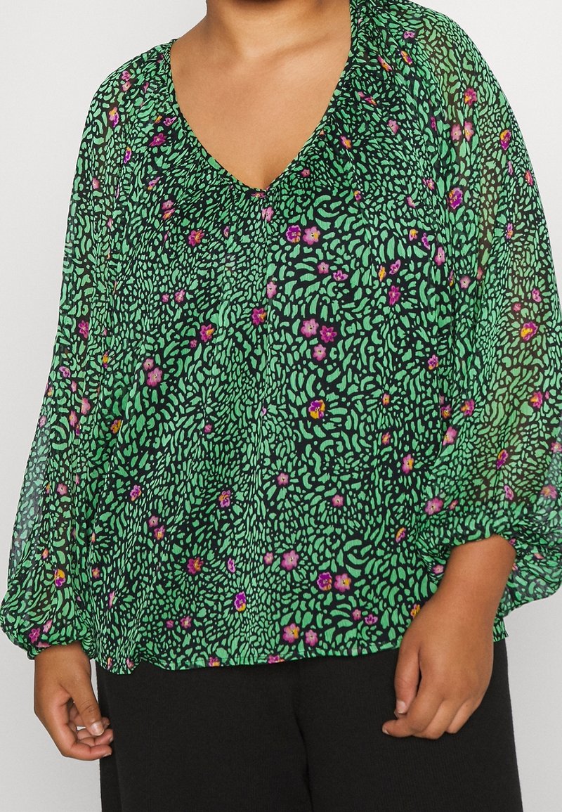Personne portant un chemisier vert avec un motif abstrait noir et de petites fleurs roses et orange, associé à un pantalon noir.