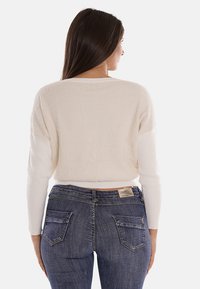 Maglione morbido di colore crema con maniche lunghe a coste, abbinato a jeans aderenti blu scuro con tasche posteriori e etichetta.