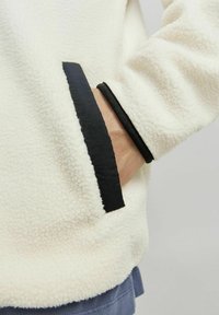 Nahaufnahme einer weißen Fleecejacke mit schwarzem Besatz. Der Stoff hat ein weiches, strukturiertes Aussehen und verfügt über ein elegantes Taschen-Detail.
