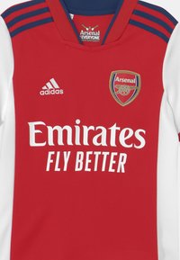Maglia da calcio rossa e bianca con colletto rotondo, con strisce blu navy sulle spalle, logo "Emirates" in bianco e stemma dell'Arsenal.