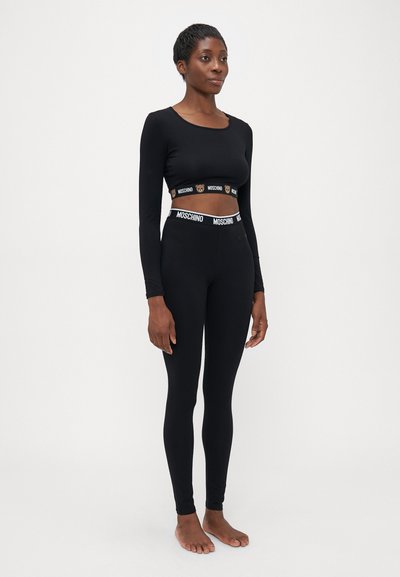 Naine seisab paljajalu, kandes musta pika varrukaga lühikest topi ja legginsid, millel on brändi Moschino elastsed vööd ja karu detail.