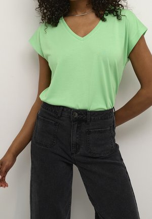 Kobieta ubrana w jasnooliwkową bluzkę z krótkim rękawem i dekoltem w serek, włożoną do czarnych dżinsów z wysokim stanem, stojąca na tle białej ściany.