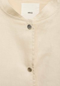 Beżowa lniana koszula z okrągłym dekoltem i dwiema widocznymi srebrnymi zatrzaskami, metka marki "MNG" wewnątrz kołnierza.