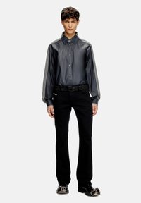 Camisa negra de botones con mangas a rayas, con una textura brillante y un ajuste relajado. Combinada con pantalones negros y calzado distintivo.