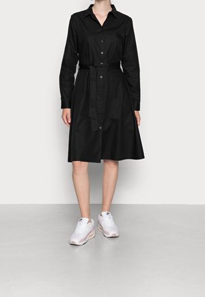 Kobieta nosząca czarną sukienkę z guzikami do kolan z długimi rękawami i paskiem w talii, w połączeniu z białymi sneakersami Nike.