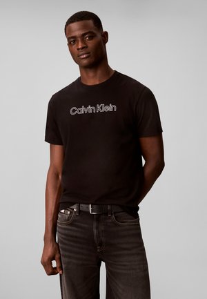 Mężczyzna ubrany w czarną koszulkę Calvin Klein i ciemne jeansy stoi na jednolitym jasnoszarym tle.