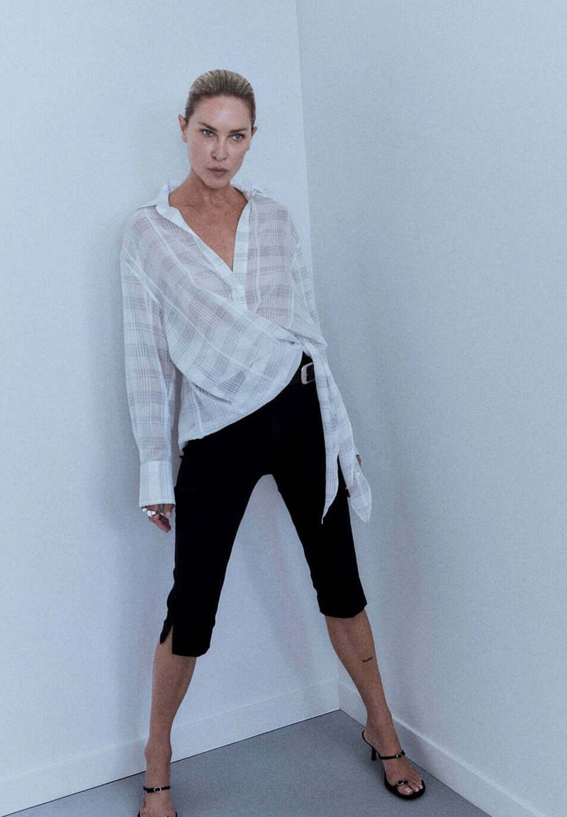 Femme aux cheveux tirés en arrière, debout contre un mur blanc d’angle, portant une chemise ample blanche à carreaux, un pantalon court noir et des sandales noires à talons.