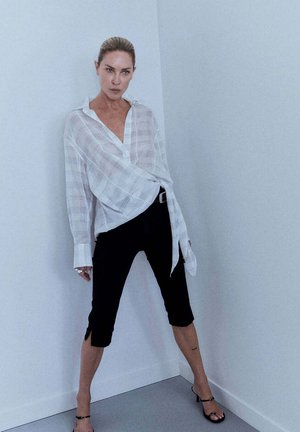 Femme aux cheveux tirés en arrière, debout contre un mur blanc d’angle, portant une chemise ample blanche à carreaux, un pantalon court noir et des sandales noires à talons.