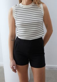 Kobieta w białej, bezrękawnikowej, prążkowanej bluzce z czarnymi poziomymi paskami i czarnych szortach z wysokim stanem, stojąca w pomieszczeniu.