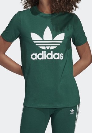 Osoba ubrana w zieloną koszulkę Adidas z białym logo oraz pasujące zielone spodnie Adidas z białymi paskami po bokach.