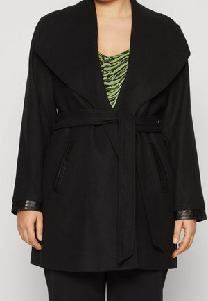 Personne portant un manteau noir ceinturé avec un large col, par-dessus un haut vert et noir à motifs, debout devant un fond clair uni.