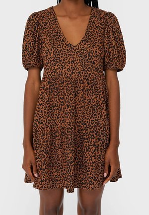 Vrouw draagt een korte bruine jurk met zwarte luipaardprint, opgeblazen korte mouwen en een V-hals, staand tegen een effen achtergrond.