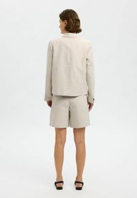 Blazer in lino beige chiaro abbinato a pantaloni corti coordinati. Il blazer presenta una singola apertura sul retro e bottoni sui polsini. Texture liscia.