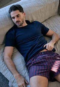 Camiseta de algodón azul marino y pantalones cortos de cuadros rojos, con cinturilla elástica, llevados por un hombre relajándose en un sofá acolchado de color gris claro.