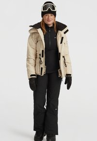 Beige pufferjack met een gestructureerd patroon, zwarte accenten en verstelbare trekkoorden, gecombineerd met een zwarte thermotop, broek en handschoenen.
