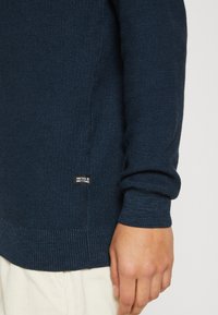 Suéter de punto azul marino con una tela texturizada, puños acanalados y una etiqueta con el logo en el lado inferior. Mano visible descansando cerca.