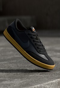 Sapatilhas de couro pretas com sola de borracha amarela, apresentando um bico redondo, atacadores planos e um logo da Nike na língua e na lateral.