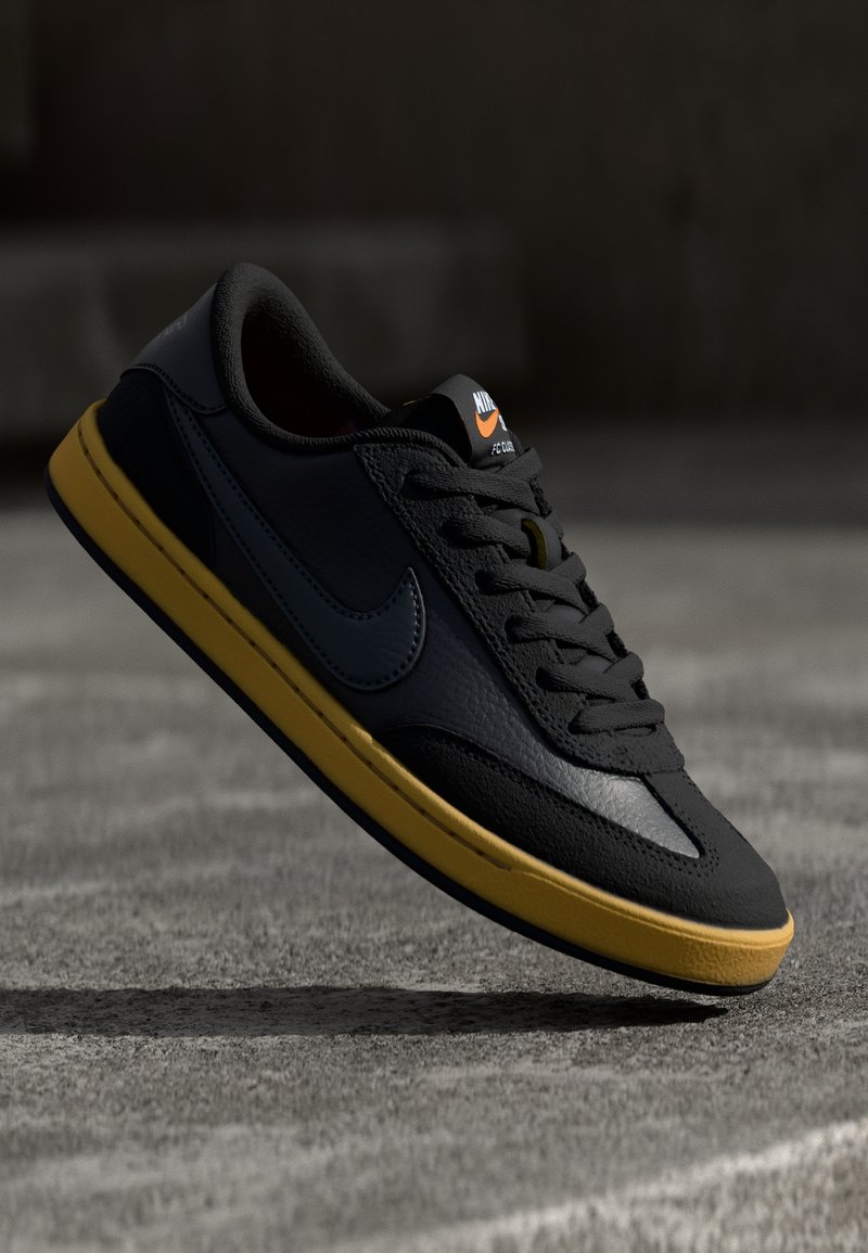 Sapatilhas de couro pretas com sola de borracha amarela, apresentando um bico redondo, atacadores planos e um logo da Nike na língua e na lateral.