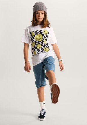 Barn iført en grå beanie, hvid t-shirt med gule smiley-ansigter, denimshorts, hvide sokker og sorte sneakers, der løfter det ene ben fremad.