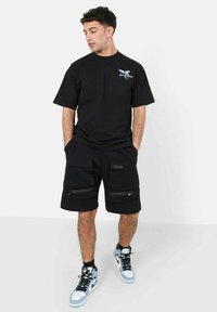 Zwarte katoenen t-shirt met een grafische print op de borst. Gecombineerd met zwarte cargoshorts, voorzien van meerdere met rits gesloten zakken en een losse pasvorm.